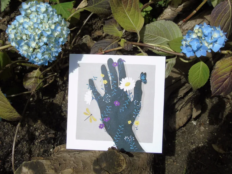 Stampa illustrata di una mano scura avvolta da fiori e farfalle, posizionata su un tronco in un giardino con ortensie blu