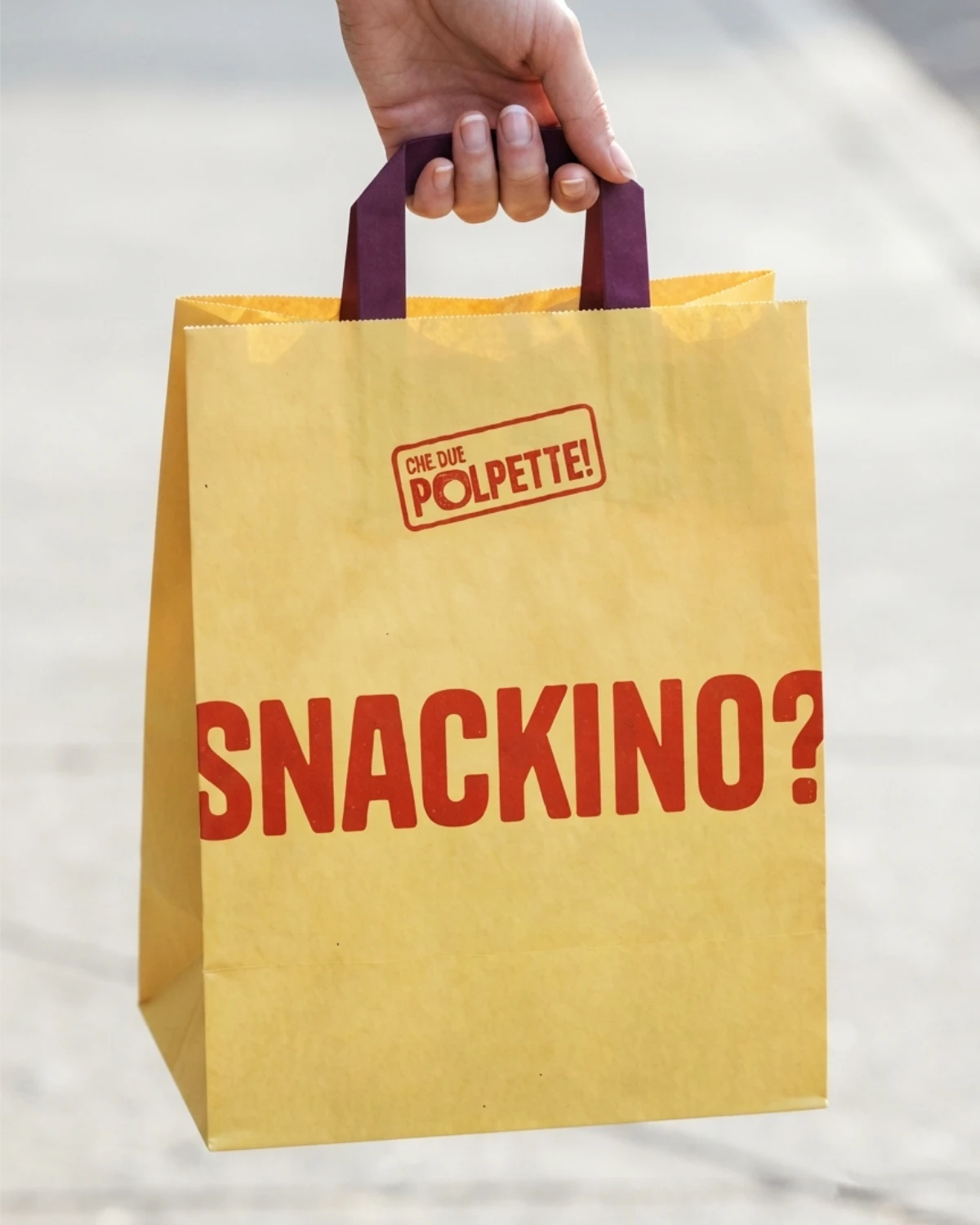 Sacchetto di carta per l'asporto con i manici, il logo e una scritta "Snackino?" stampata, che mette in evidenza il tono di voce informale del brand.