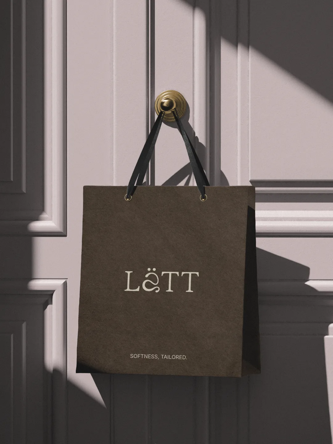 Busta per lo shopping marrone con il logo e il motto Lätt, appesa alla maniglia dorata di una porta rosa pastello.
